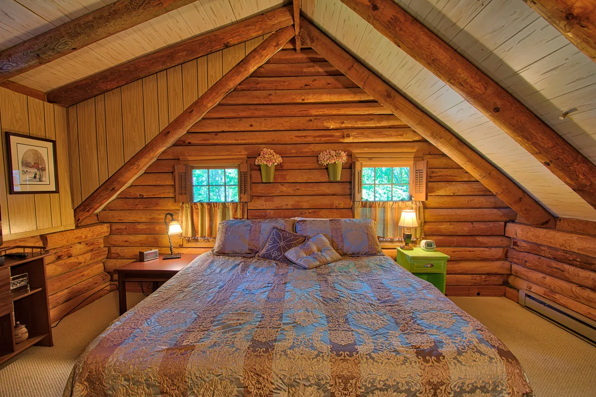 A cozy master bedroom featuring a bed with ornate bedding, log walls, a green nightstand, and windows adorned with flower pots. Sunlight filters in, hinting at views of the yard and distant peaks, while the warm wood accents create a restful ambiance.