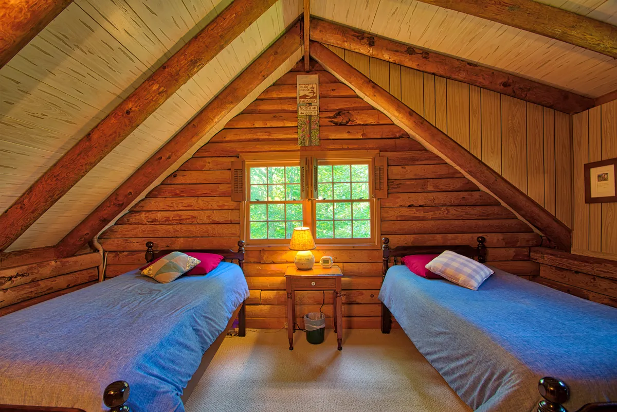 A bright, inviting bedroom with two twin beds, a wooden nightstand and lamp between them, and a window that frames lush greenery outside. The soft morning light and tranquil decor provide a welcoming space for friends or small families.