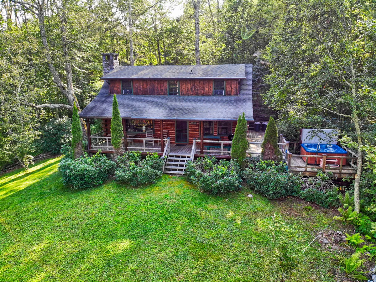 The image shows a charming log cabin nestled amid lush greenery, with a spacious deck featuring a bubbling hot tub. The scene evokes tranquility, with the golden glow of sunset casting warm light over the inviting outdoor spaces—ideal for relaxing soaks, stargazing, and gatherings with loved ones.