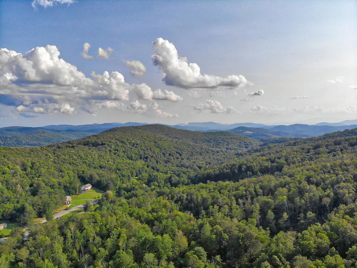 A sweeping view of rolling hills, dense forests, distant mountains, and a moody sky with scattered small houses. The landscape evokes the quintessential Blue Ridge vistas, inviting couples to explore nearby trails, enjoy sunsets, or simply marvel at the mountain beauty together.