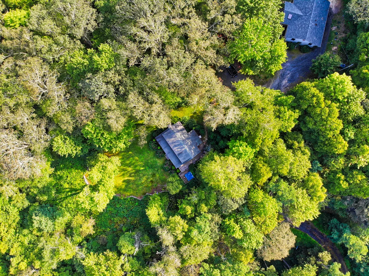 This image captures the exterior of Winds Aloft, showing the house nestled among trees and greenery. The inviting hot tub is positioned on the deck, with the surrounding lawn and nature trails visible. The porch offers a perfect vantage point for morning coffee or evening relaxation, tying directly to the section’s emphasis on romance and scenic views.