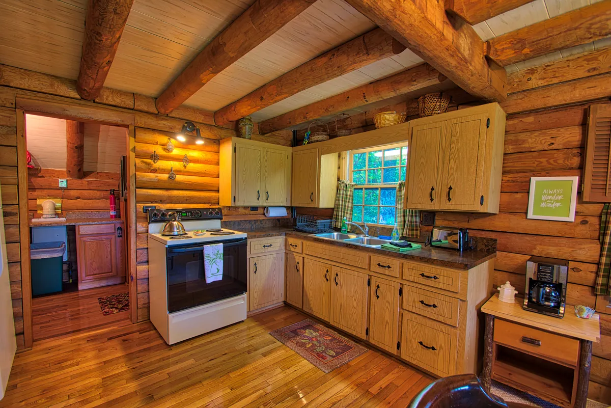 A charming, fully equipped kitchen with wooden cabinets, a stove, and a sunlit window. The space feels homely and functional, ideal for preparing home-cooked meals or morning coffee rituals, with wooden beams overhead adding to the cabin’s rustic character.