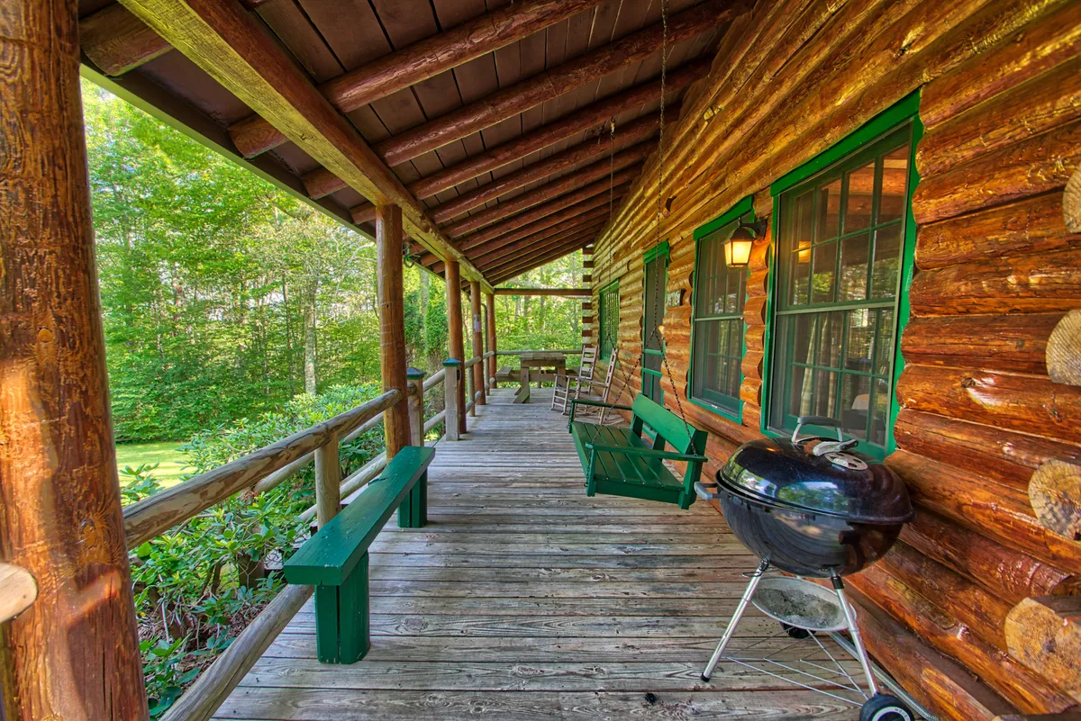 This exterior image highlights the log cabin's porch area, featuring green benches, a grill, and swinging chairs. The cozy setup suggests relaxation and social gatherings, with visible seating options ideal for rocking chairs and porch swings.