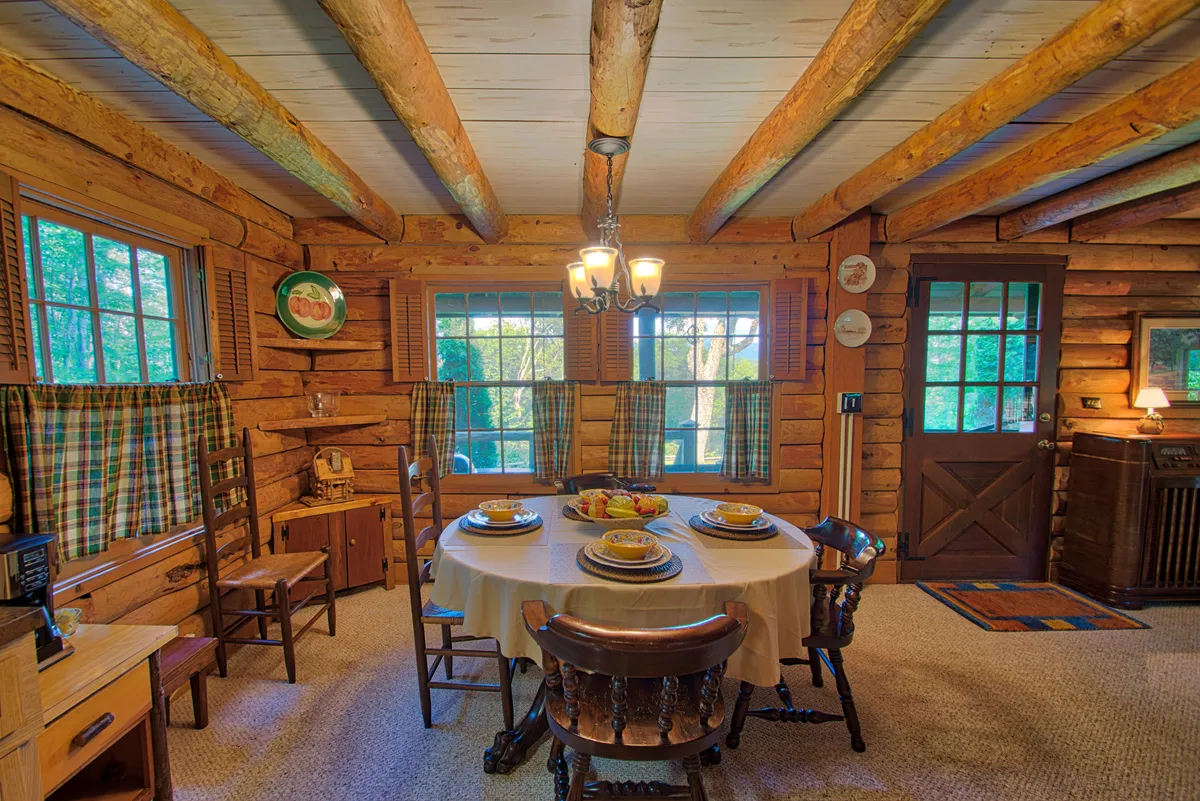 A welcoming dining area with a wooden table set for a meal, surrounded by warm wood tones, exposed beams, and decorative plates. Windows with curtains frame the space, allowing natural light to filter in, and creating an inviting setting for family dinners or friendly gatherings.