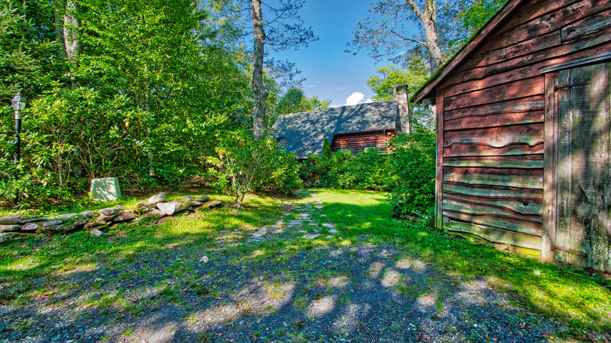 This exterior image shows a rustic cabin with a weathered shed, enveloped by lush greenery and featuring a prominent stone pathway leading up to the entry. The foreground is defined by the stone path, perfectly echoing the rhododendron-lined approach described in the content. The overall mood is tranquil and inviting, setting the stage for an adventure or welcome home after outdoor activities.