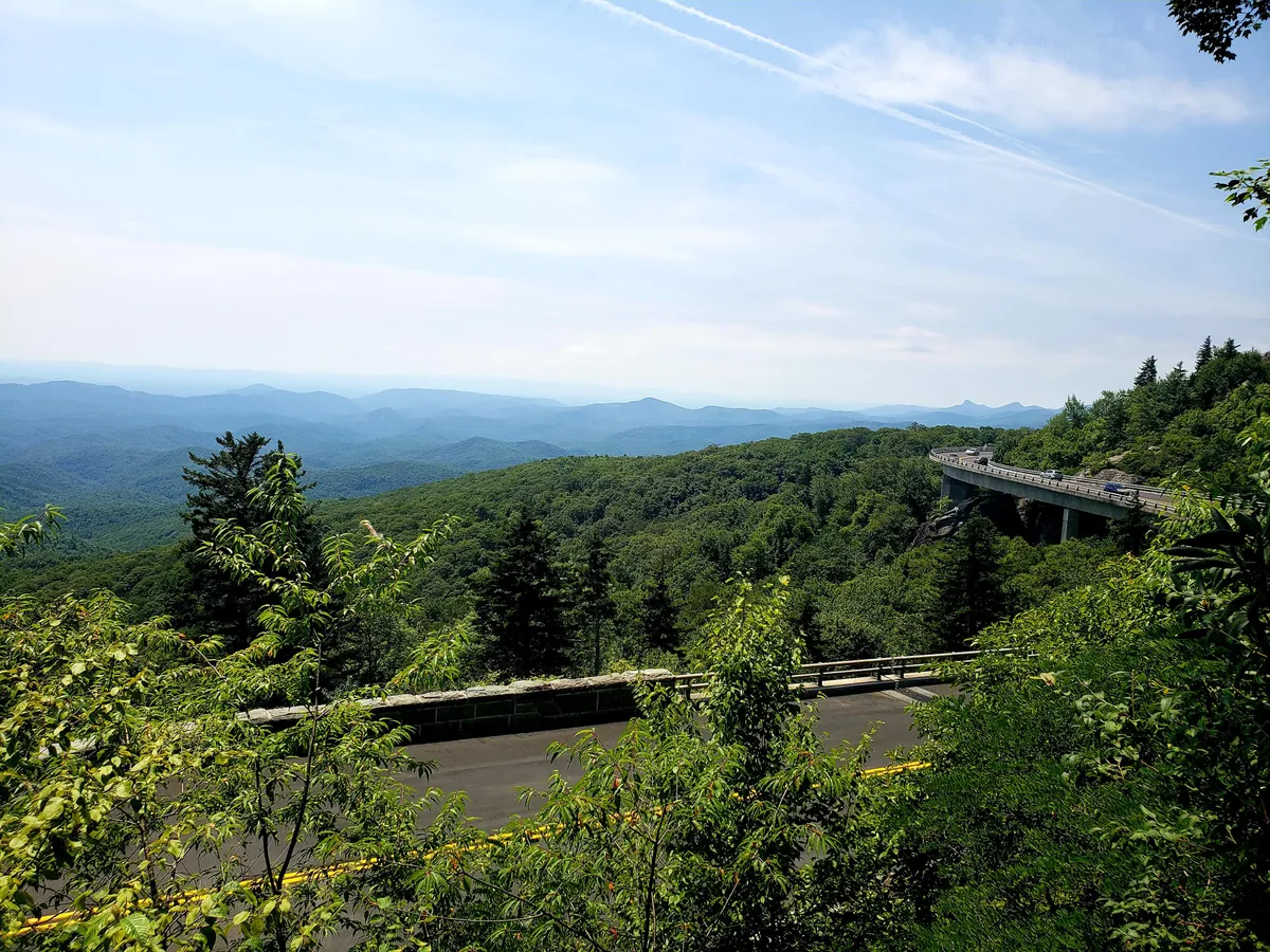 A classic Blue Ridge Parkway scene, featuring sweeping mountain ranges, a winding road traversing lush greenery, and an expansive sky. The landscape invites scenic drives and adventure, capturing the allure of nearby attractions like Grandfather Mountain and the Linn Cove Viaduct.