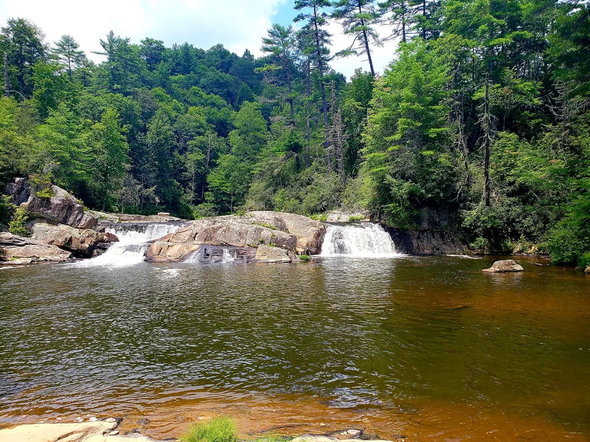 A scenic waterfall surrounded by forest greenery and dramatic rock formations. The scene evokes the natural wonders waiting to be explored, inviting viewers to imagine hiking, picnicking, or capturing photos in this serene mountain environment.