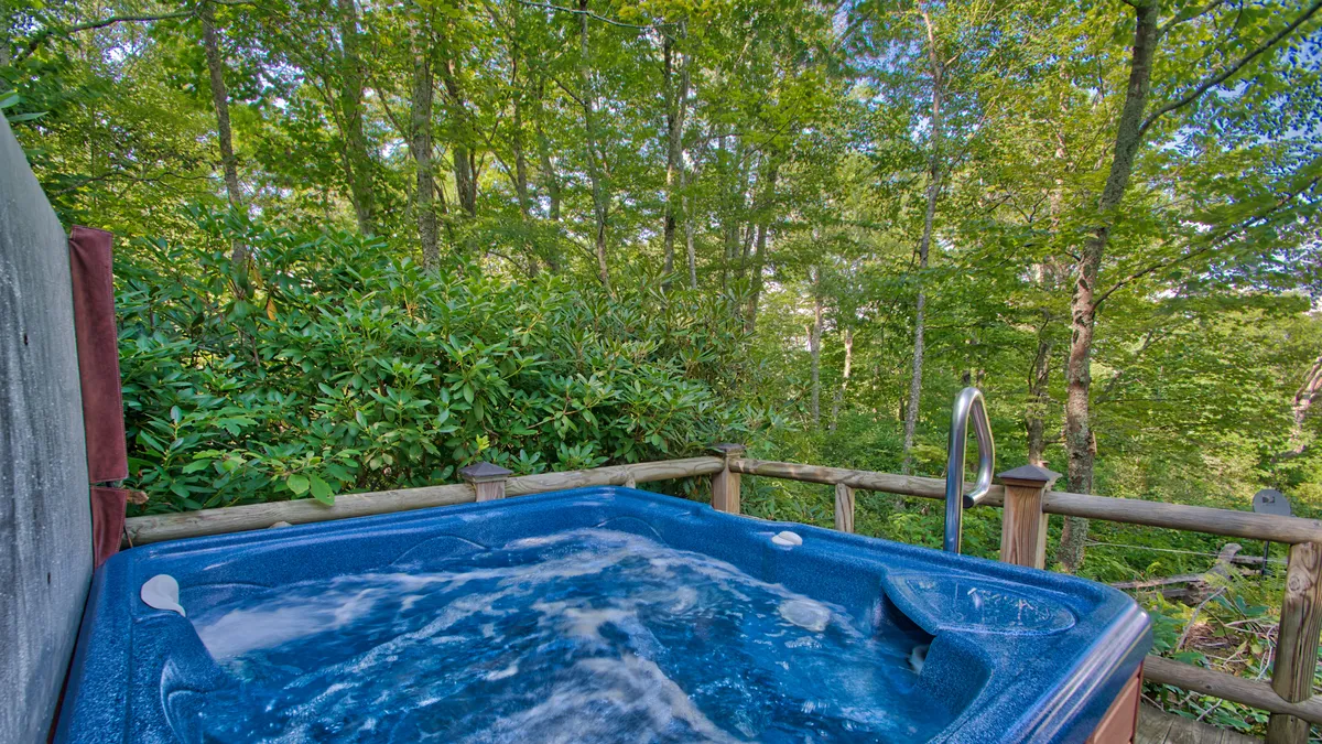 A tranquil outdoor scene focusing on the hot tub set against a backdrop of lush greenery and a sturdy wooden railing. The hot tub feels private yet connected to the surrounding landscape, inviting guests to soak while enjoying mountain views.