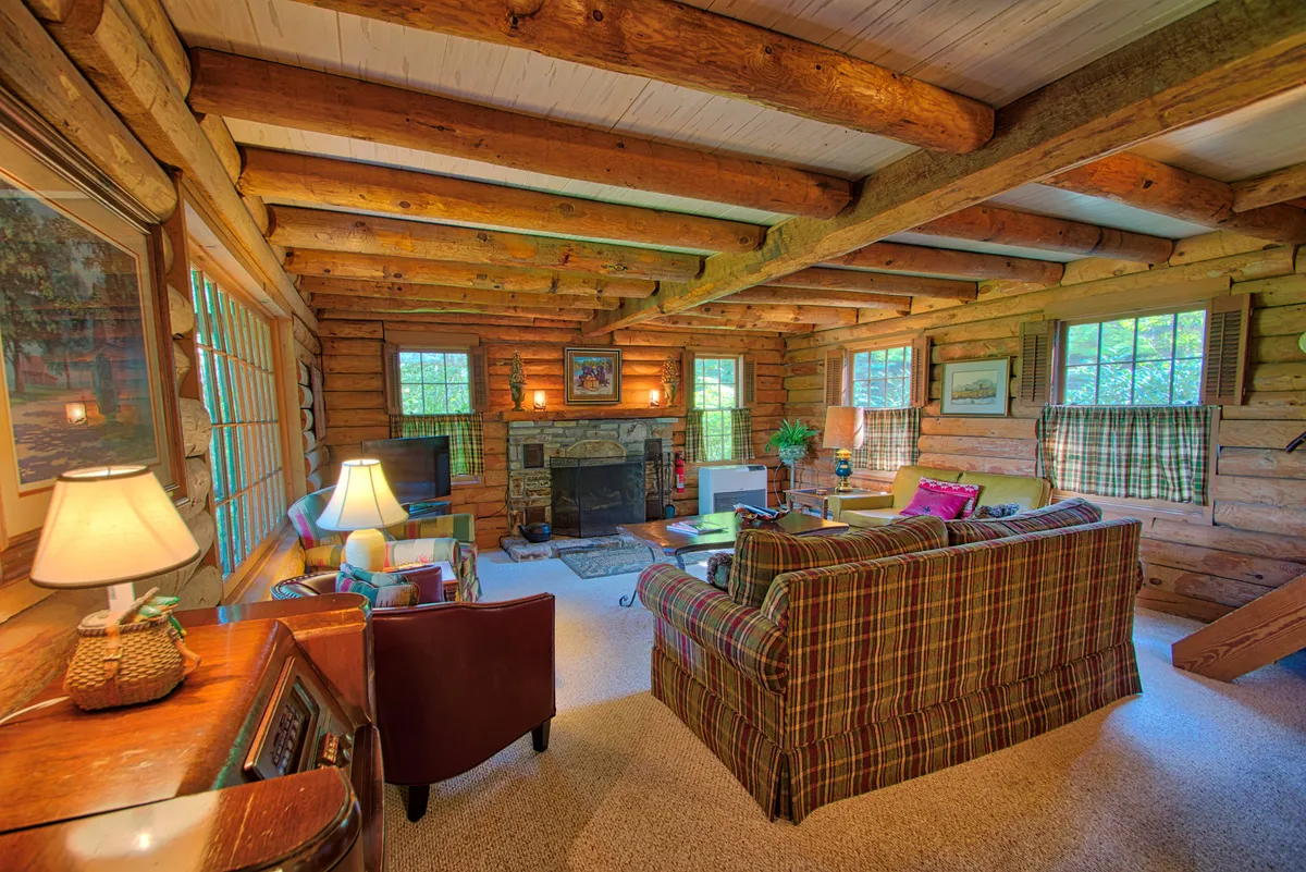 A warmly lit living room featuring a striking stone fireplace as the centerpiece. The space includes a cozy plaid sofa, exposed wooden beams, and large windows that let in natural light, offering glimpses of the mountain views beyond. The inviting layout encourages gathering by the fire, relaxing with a book, or enjoying time with loved ones.